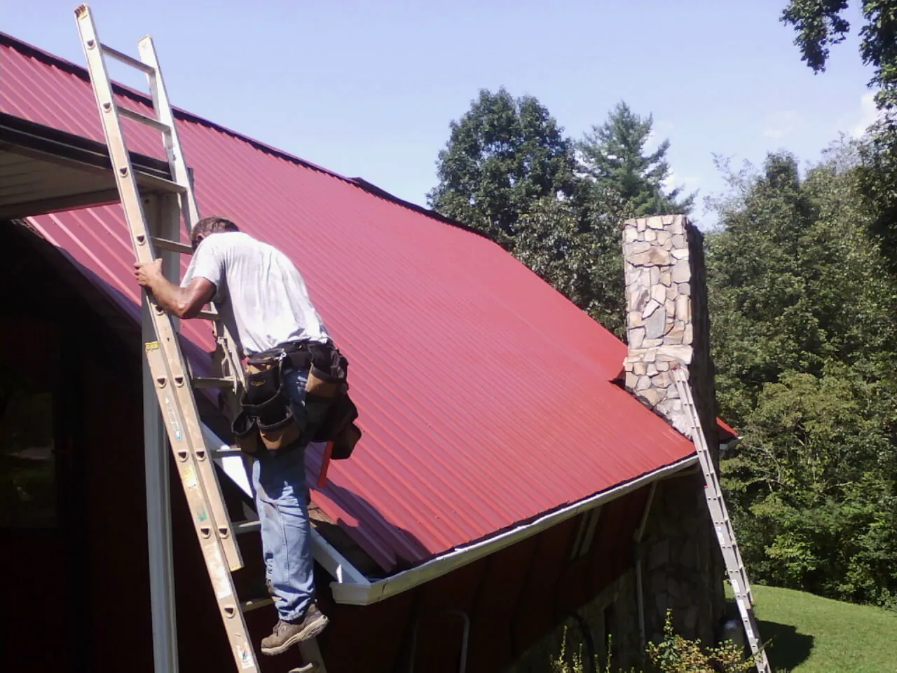 Roofer on a ladder beside a red metal roof and stone chimney — Soffit & Fascia Repair by Roofing Brothers in Afton